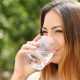 Happy healthy woman drinking fresh water from a glass outdoor with a green background