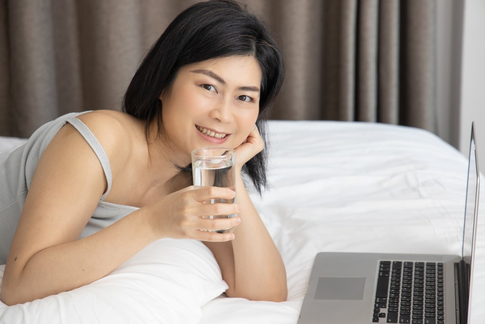 women drinking water on bed while using laptop